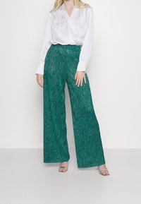 Tyrkysové široké kalhoty vyrobené z texturované tkaniny s kovovými akcenty, kombinované s bílou zavinovací blůzou. Průhledné sandály na podpatku.