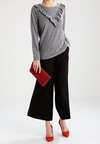 Femme portant un pull gris à manches longues avec des volants, un pantalon noir à jambes larges, des escarpins rouges à bouts pointus, et tenant une pochette rouge.