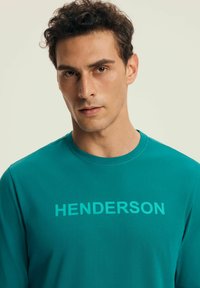 Turkusowa koszulka z długim rękawem wykonana z miękkiego materiału, z napisem "HENDERSON" nadrukowanym na klatce piersiowej w kontrastowym kolorze.