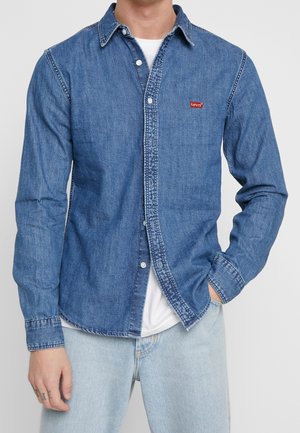 Mężczyzna w niebieskiej, dżinsowej koszuli Levi's zapinanej na guziki, na białym t-shircie i jasno-niebieskich dżinsach, z jedną ręką w kieszeni, na jednolitym tle.