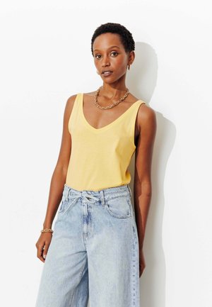 Femme aux cheveux courts portant un haut jaune sans manches, un jean taille haute bleu clair, un collier chaîne doré et un bracelet, debout contre un mur blanc.