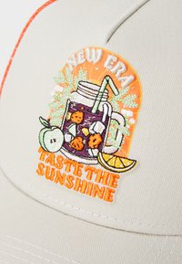 Beige kšiltovka s vyšitým designem zobrazujícím sklenici s nápojem, ovoce a text "Ochutnej slunce" v oranžové barvě. Texturovaná tkanina a oranžové akcenty.