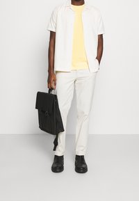 Camicia a maniche corte di colore chiaro, t-shirt gialla, pantaloni beige, zaino nero con pattina e stivaletti neri alla caviglia. Design e trame semplici.