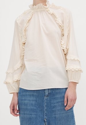 Kobieta ubrana w kremową bluzkę z długim rękawem ozdobioną falbankami oraz niebieską spódnicę dżinsową, stojąca z rękami swobodnie opuszczonymi wzdłuż ciała.