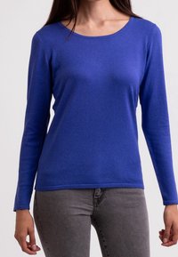 Blauwe langarm gebreide top met een ronde halslijn en een gladde textuur, gecombineerd met grijze jeans. Eenvoudig ontwerp zonder zichtbare patronen of accenten.