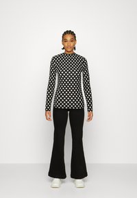 Blusa preta de mangas longas com bolinhas brancas, apresentando um decote alto. Combinada com calças pretas largas e ténis brancos.