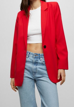 Frau trägt einen roten Blazer über einem weißen Cropped-Top und hellblauen, hoch taillierten Jeans vor einem einfarbigen Hintergrund.