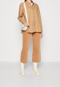 Camisa marrón de botones, vaqueros anchos color beige, botas de tobillo color crema y un pequeño bolso cruzado blanco. Texturas suaves y diseño casual.