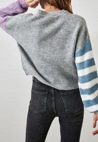 Grauer gerippter Pullover mit lila und blau gestreiften Ärmeln, kombiniert mit schwarzer Jeans aus Denim. Fokus auf Textur und kontrastierende Farben.