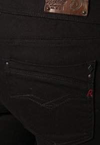 Poche arrière d'un jean en denim noir avec des coutures décoratives, de petits rivets, une lettre R rouge brodée, et un patch en cuir marron au-dessus.