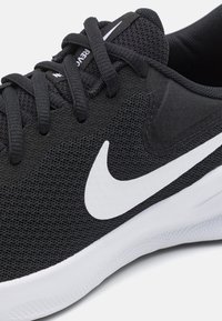 Svart sportskor med nätöverdel, vitt Nike swoosh-logotyp, runda skosnören och en vit gummisula med ett texturerat mönster.