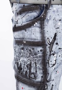 Jeans in denim sbiadito con design a macchie di vernice in bianco e nero, dotati di molteplici tasche e dettagli di cucitura visibili.