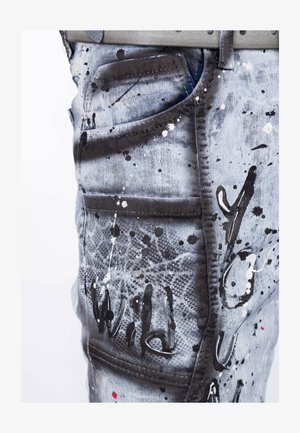 Faded denim jeans with splatter paint design in black and white, featuring multiple pockets and visible stitching details.