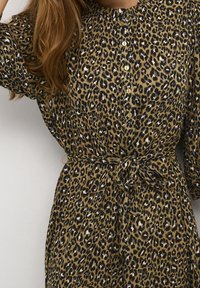 Šaty s leopardím potiskem v tan a černé barvě, s knoflíkovým zapínáním vpředu, krátkými rukávy a detailem vázání v pase. Texturovaný povrch.