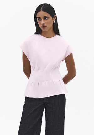 Kobieta o długich ciemnych włosach nosi jasnoróżowy top bez rękawów z zakładkami w talii oraz czarne spodnie o szerokich nogawkach, stoi z rękami za plecami.