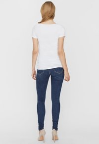 Witte kortebloes met korte mouwen gecombineerd met donkerblauwe skinny jeans, met een gladde textuur en subtiele stiknaden op de zakken, gedragen met beige hakken.