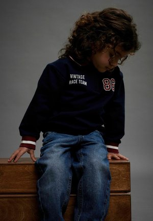 Granatowy sweter z napisem "VINTAGE RACE TEAM" i numerem 86, w połączeniu z niebieskimi dżinsami. Sweter ma czerwono-białe paski na mankietach. Pozycja siedząca.