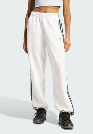 Kobieta w białych spodniach dresowych Adidas z czarnymi paskami po bokach i czarnych butach sportowych, stojąca na neutralnym tle.