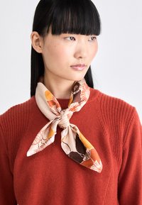 Pañuelo de seda en color crema con patrones florales en naranja y marrón, atado en forma de lazo alrededor del cuello, usado sobre un suéter rojo texturizado.