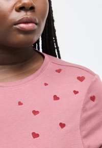 Primo piano di una persona che indossa una camicia rosa con stampe di cuori rossi sparse sulla spalla e sul petto.