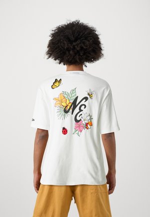 Person med krøllet hår iført en hvid t-shirt med farverige blomster, insekter og bogstaver "NE" på ryggen, kombineret med sennepsgule shorts.