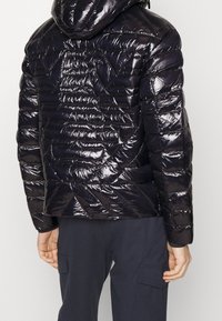 Černá, lesklá péřovka s kapucí, zdobená prošívanými diamantovými vzory a elastickými manžetami, navržená pro teplo a přiléhavý vzhled.