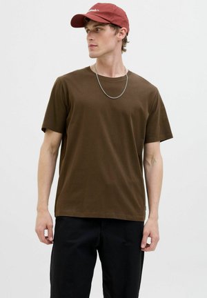 T-shirt en coton marron à manches courtes, col rond et coupe décontractée, associé à une casquette de baseball rouge et un collier en chaîne en argent.