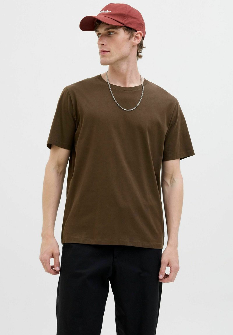 Bruine katoenen T-shirt met korte mouwen, ronde halslijn en losse pasvorm, gecombineerd met een rode baseballpet en een zilveren ketting.
