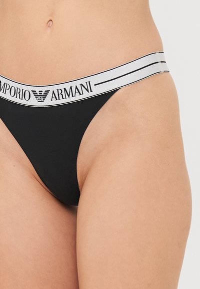 Black thong with a soft elastic waistband featuring white text and stripes. The material appears smooth with a minimalistic design.