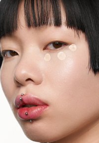 Närbild av ett ansikte med slät, ljus hy; tre ljusbeige prickar av concealer under ena ögat; genomborrade läppar med metallstift.