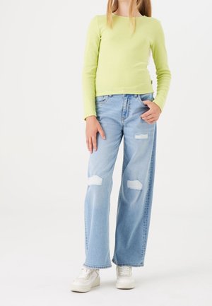 Dziewczyna nosząca jasnozieloną bluzkę z długim rękawem, jasnoniebieskie podarte szerokie dżinsy i białe trampki, stoi z jedną ręką w kieszeni.