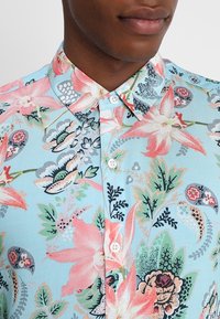 Chemise courte à manches courtes bleu clair avec un motif floral vibrant en rose, vert et noir. Dotée d'un col à boutons et de boutons sur le devant.