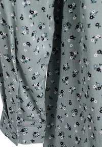 Blusa verde con un motivo floreale caratterizzato da fiori neri, bianchi e blu. Il tessuto ha una finitura testurizzata e polsini con bottoni.