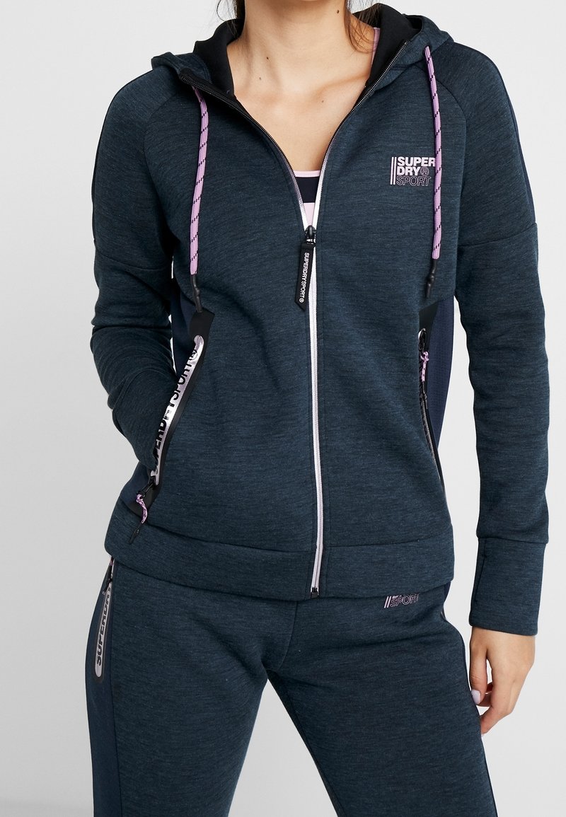 Sudadera con cremallera de color azul marino con cordones rosas y una marca. Presenta bolsillos laterales y acentos de material en contraste en las mangas.