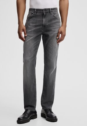 Mann trägt gerade geschnittene graue Jeans und schwarze Leder-Schnürschuhe, steht vor einem schlichten hellen Hintergrund.