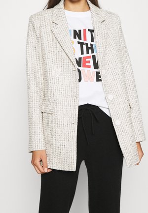 Texturerad krämfärgad blazer med en strukturerad design, slagkragar och två framknappar, bärs över en grafisk vit t-shirt och svarta byxor.