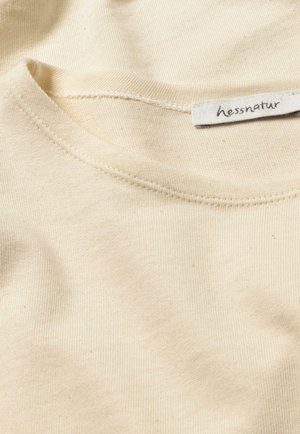Dettaglio di una T-shirt di cotone color crema con scollatura a costine e cuciture visibili. L'etichetta recita "hessnatur". Tessuto morbido senza motivi.