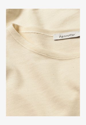 Cremfarbendes Baumwoll-T-Shirt-Detail mit ribbeltem Ausschnitt und sichtbaren Nähten. Das Etikett trägt die Aufschrift "hessnatur". Weiche Textur ohne Muster.