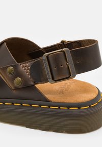 Bruine leren sandaal met een gestructureerde strap, een verstelbare metalen gesp en een gevoerde, tan kleurige binnenzool, met gele stiksels op een dikke zwarte zool.