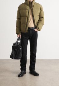 Giacca imbottita verde oliva, maglione beige, jeans neri e stivali in pelle nera. Tiene una borsa in pelle nera con un design arrotondato.