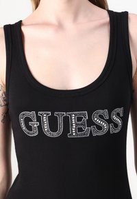 Top de alças preto com nervuras, apresentando "GUESS" em strass prateados ao longo do peito. Decote em barco e design ajustado.