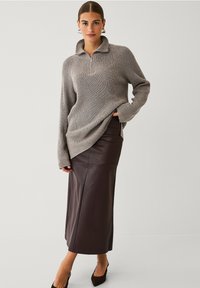 Kvindelig iført en grå ribbet sweater med halv lynlås og en brun læder midi-nederdel, stående foran en ensfarvet lys baggrund.
