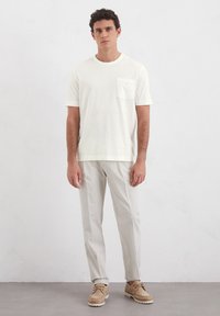Helles Cremefarbiges T-Shirt mit kurzen Ärmeln und einer Tasche, kombiniert mit hellgrauen tapered Pants und beigen Schuhen. Der Stoff wirkt weich und strukturiert.