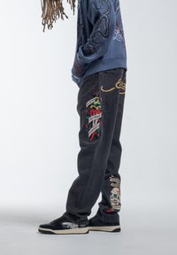 Zwarte denim jeans met kleurrijke geborduurde patches met zinnen en ontwerpen, een relaxed fit en gedetailleerde accentstiksels.