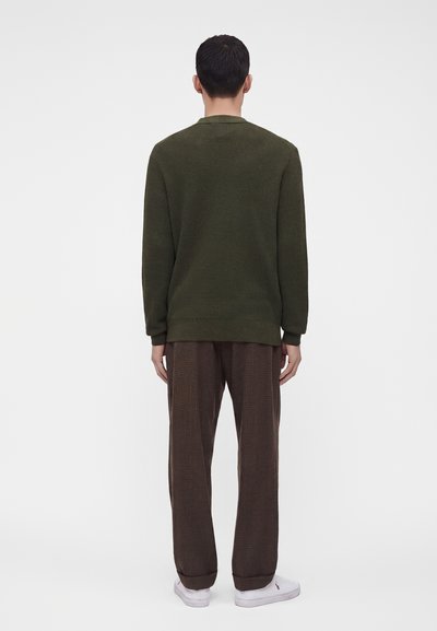 Homme portant un pull texturé vert foncé, un pantalon à carreaux marron et des baskets blanches, debout de dos devant un fond clair uni.