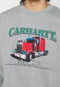 Šedá mikina se grafikou červeného kamionu, s textem "CARHARTT" v zelené barvě nahoře a "WORK IN PROGRESS" dole.
