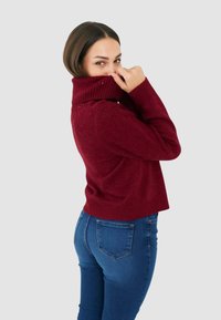 Maglione dolcevita color bordeaux con polsini e orlo a coste, decorazioni sottili sulle spalle, abbinato a jeans blu aderenti.
