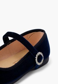 Bailarina de terciopelo azul marino con punta redondeada, que presenta una pequeña correa y un detalle de hebilla circular de rhinestones en el costado.