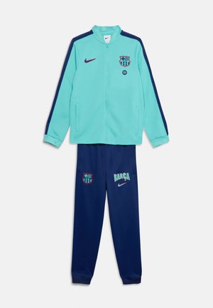 Veste zippée bleu clair avec des rayures bleu foncé et l'écusson du FC Barcelone, associée à un pantalon bleu foncé avec le texte Barça et l'écusson assorti.
