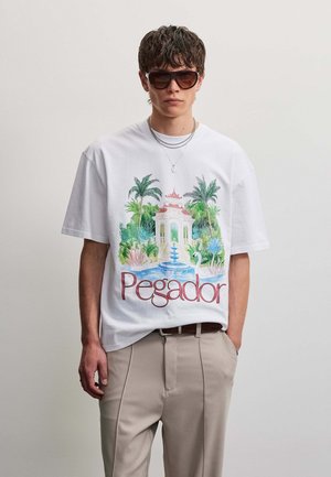 Ung mand iført en oversized hvid T-shirt med tropisk have- og fontanetryk og ordet "Pegador", beige bukser og mørke solbriller.